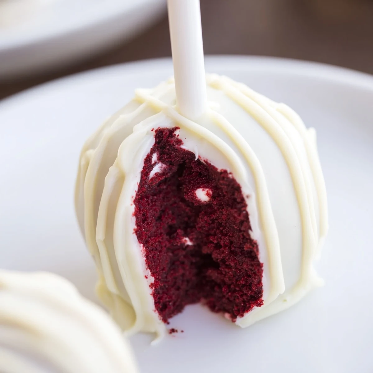 Red velvet cake pops with white chocolate drizzle stand on a serving platter, showcasing their glossy coating and moist crumbs.  