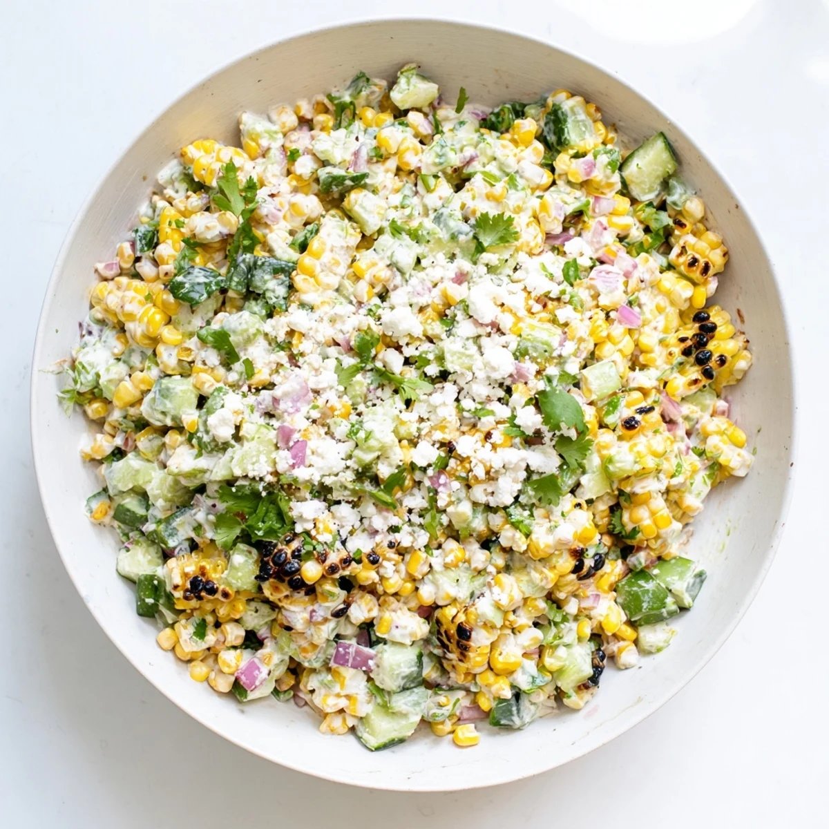 Creamy Mexican street corn salad in white bowl with diced cucumbers and red onion
