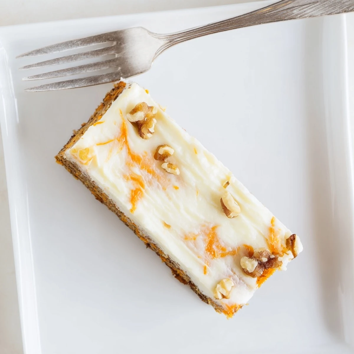 Golden carrot cake bars with swirled cream cheese layer topped with chopped walnuts on a white serving plate